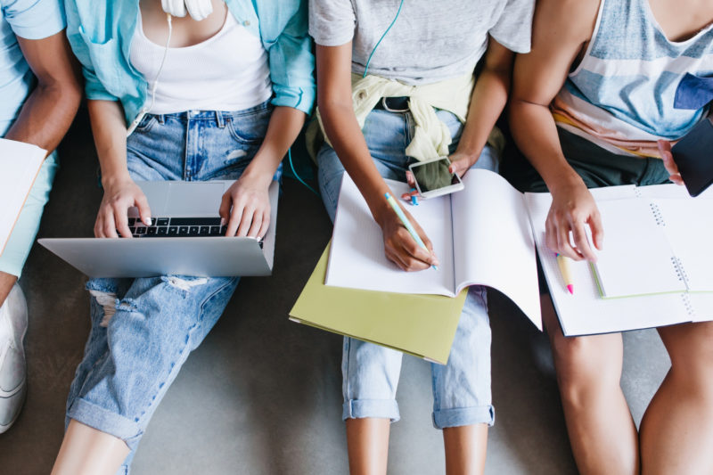 close up overhead portrait girl blue shirt jeans holding laptop knees while sitting beside university mates female student writing lecture notebook using phone friends